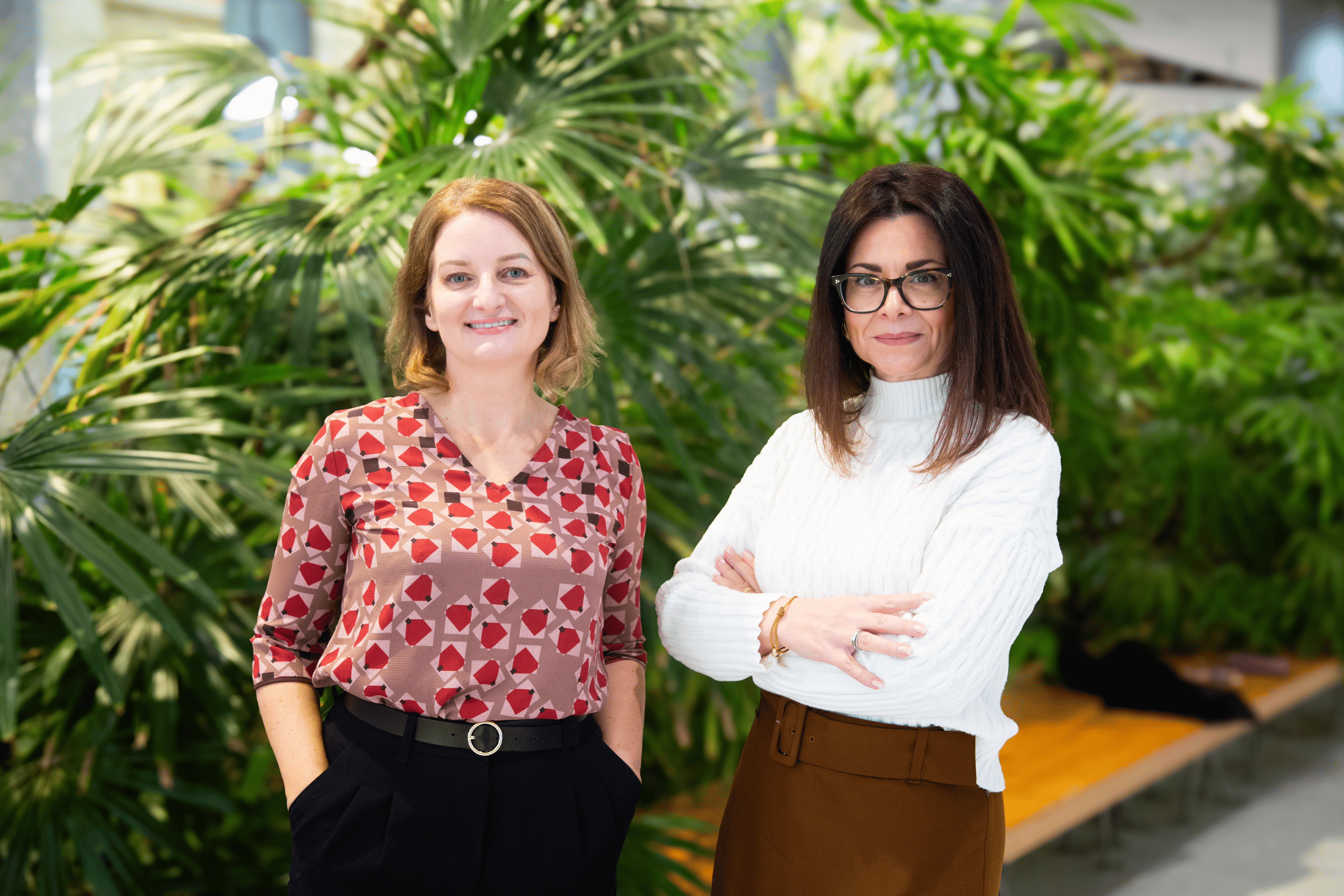 Two women pose confidently for a picture in front of lush green vegetation.